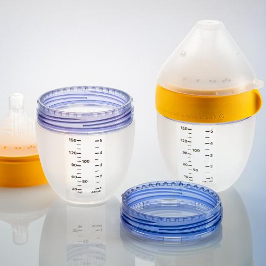 Two baby bottles and their parts on a reflective surface. One bottle is fully assembled with a yellow ring; the other is disassembled, showing a milk-filled container beside its purple and blue rings.