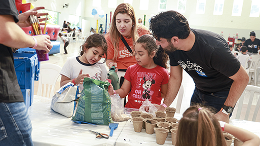 Two adults and two young girls sit around a table indoors, planting small pots with soil.