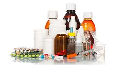 Various medications, including bottles, blister packs, loose capsules, and a syringe, on a reflective surface against a white background.
