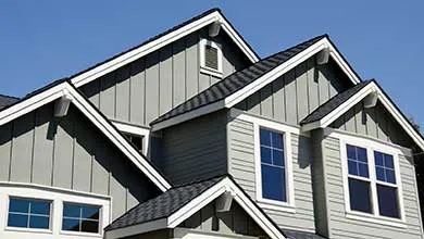 Low-angle view of a house with gray siding, dark shingled roof, and white trim, against a clear blue sky.
