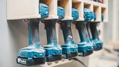 Several blue and black cordless drills hang upside down from a wooden rack.