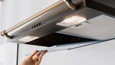 A hand opens the mesh filter on the underside of a dark kitchen range hood.