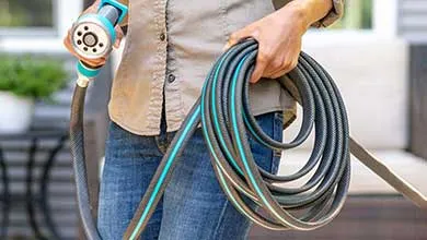 A person in a shirt and jeans holds a coiled gray and teal garden hose with a spray nozzle.