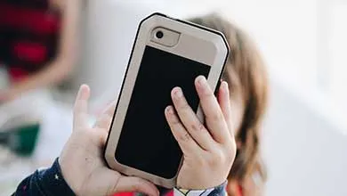 A child holds a smartphone in a beige and black case, partially covering their face.