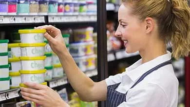 Smiling female grocery store worker in a striped apron stocking small food containers on a refrigerated shelf.