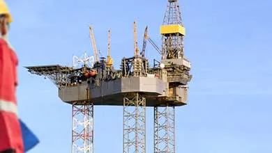 A worker wearing a hard hat and red jacket faces a large offshore oil rig with tall legs under a clear blue sky.