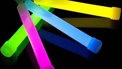Four neon glow sticks—yellow, light blue, pink, and green—are arranged in a crisscross pattern on a solid black background. Each has a visible plastic cap and hanging loop.