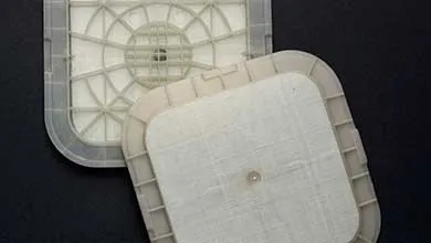 Two square plastic air filter parts with rounded corners on a dark surface; one shows a translucent grid-like frame, the other a white textured filter with a small central mounting hole.