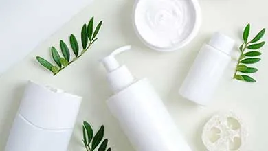 White skincare and beauty products on a light surface, including a pump bottle, open cream jar, cylindrical bottles, a loofah, and green leaf sprigs for decoration.