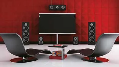 A modern home theater room with a flat-screen TV centered on a red textured wall, flanked by two tall speakers and two smaller bookshelf speakers, plus additional speakers mounted above the TV. Two black curved lounge chairs face the TV, separated by a low coffee table with books, all on white flooring.