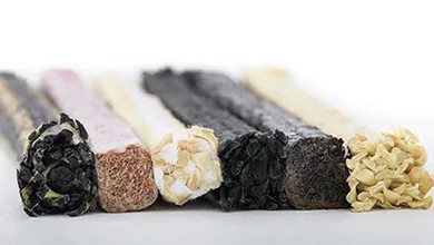Seven cylindrical snack sticks in black, lavender, white, charcoal, beige, dark gray, and golden yellow, lined up on a white background, showing their textured circular cross-sections.