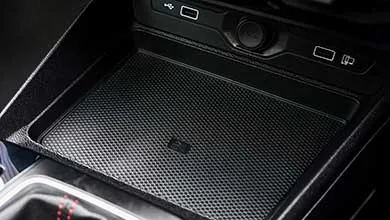 Close-up of a car center console showing a rectangular storage compartment with a textured black anti-slip mat and a wireless charging pad in the center. Above it, a dark panel houses two USB ports, a circular control dial, and additional buttons.