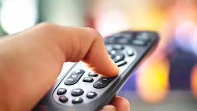 Close-up of a hand holding a black TV remote, thumb pressing a button, against a blurred warm-lit background.