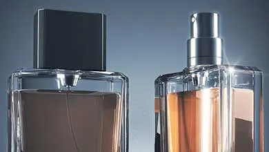 Close-up of two faceted glass cologne bottles on a blue-gray background; the left bottle has a square black cap, and the right has a silver spray nozzle.