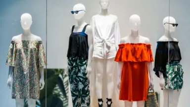 Five mannequins stand in a store window, displaying summer outfits including off-the-shoulder dresses, shorts, and leaf-print pants.